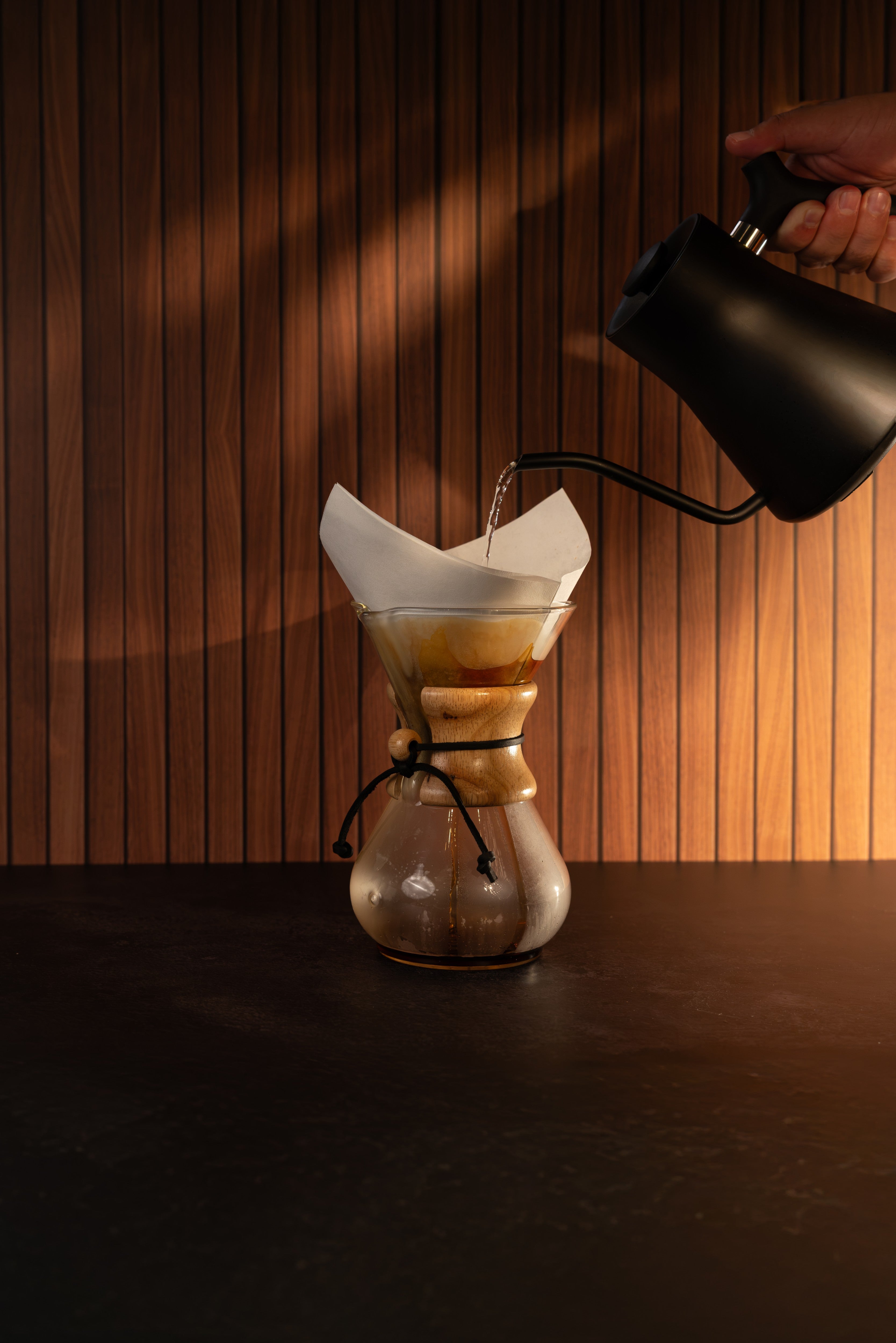 A Chemex-style pour-over coffee maker with a white paper filter and glass chamber sits on a dark tabletop, with hot water being poured from a matte black kettle against a warm wood slat wall.