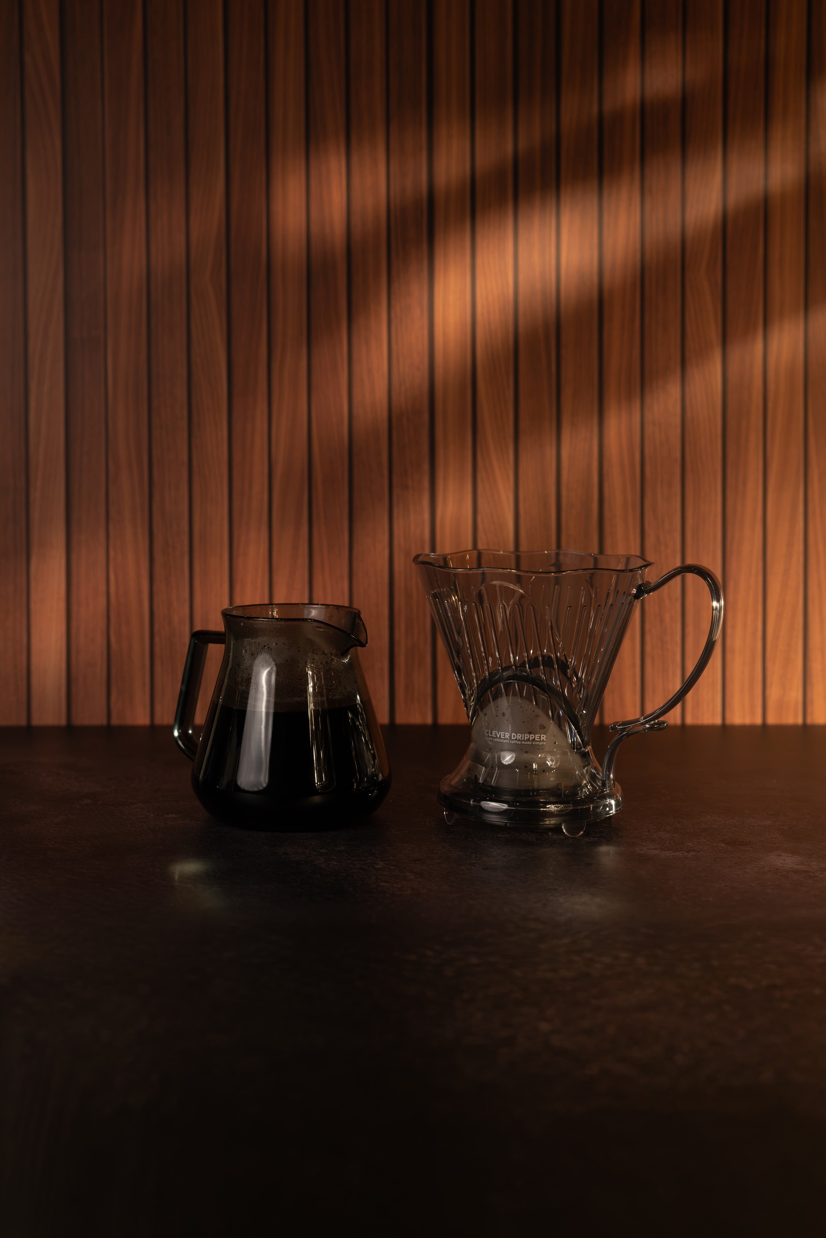 A dark glass pour-over coffee dripper and matching dark glass carafe resting on a dark wooden table against a wood-paneled wall with dramatic warm light shadows.