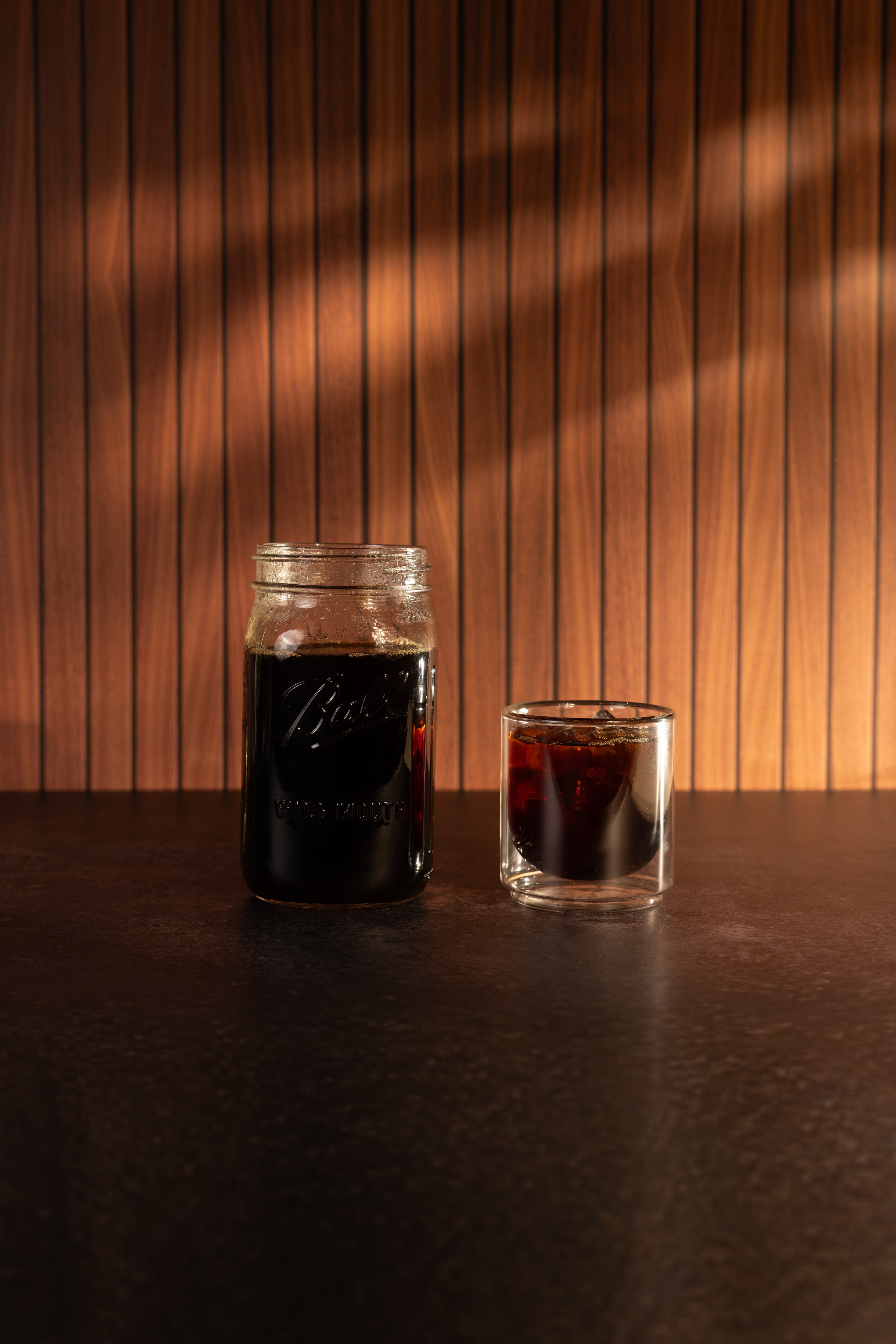 A clear glass mason jar filled with dark coffee and a small clear glass tumbler with a matching dark coffee layer, set on a dark tabletop against a warm wood-paneled wall with dramatic shadow patterns.