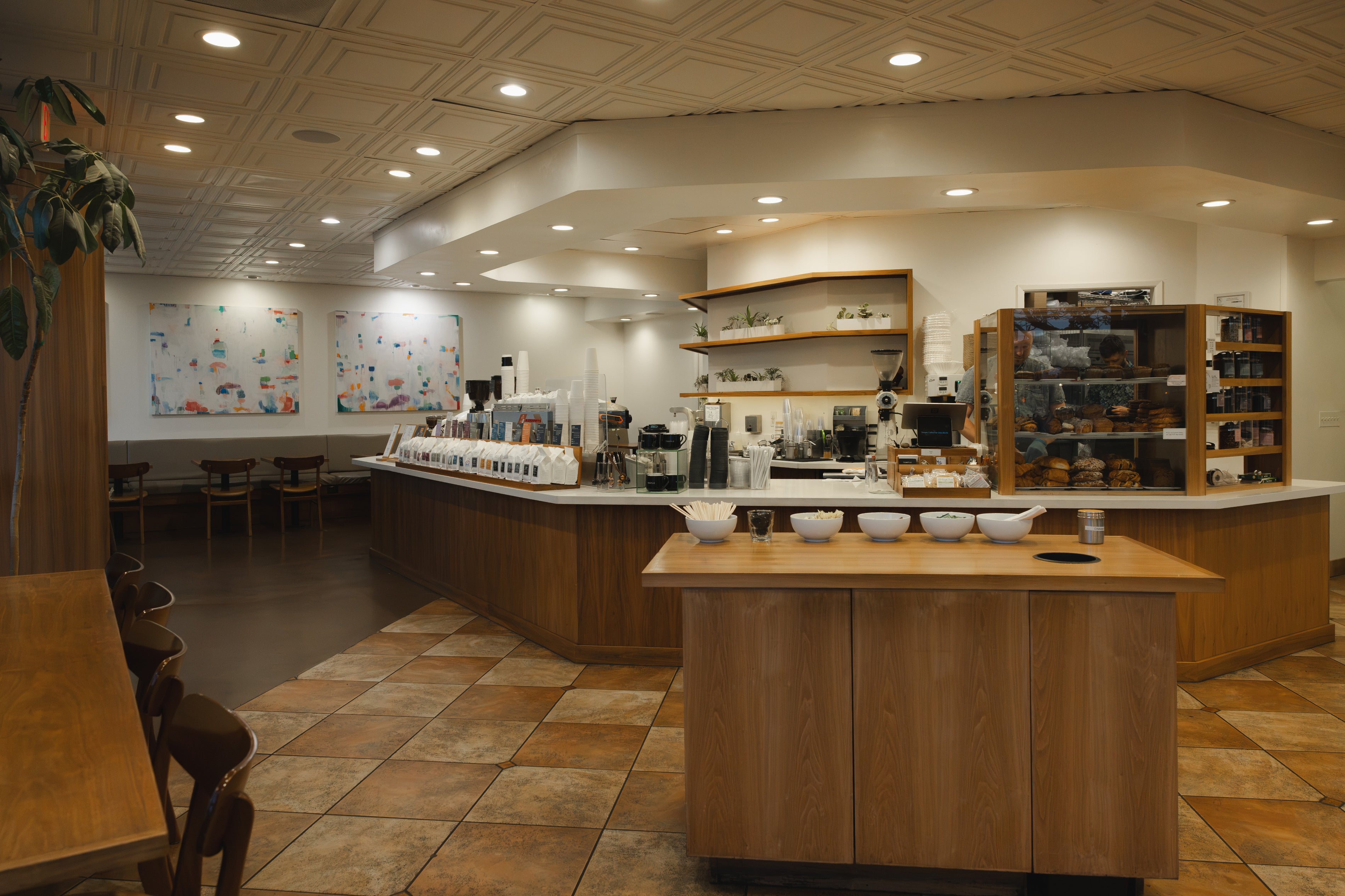 A wide interior view of a cafe or coffee shop with wooden counter islands, tiled floors, shelving, coffee equipment, and seating areas under recessed ceiling lights.