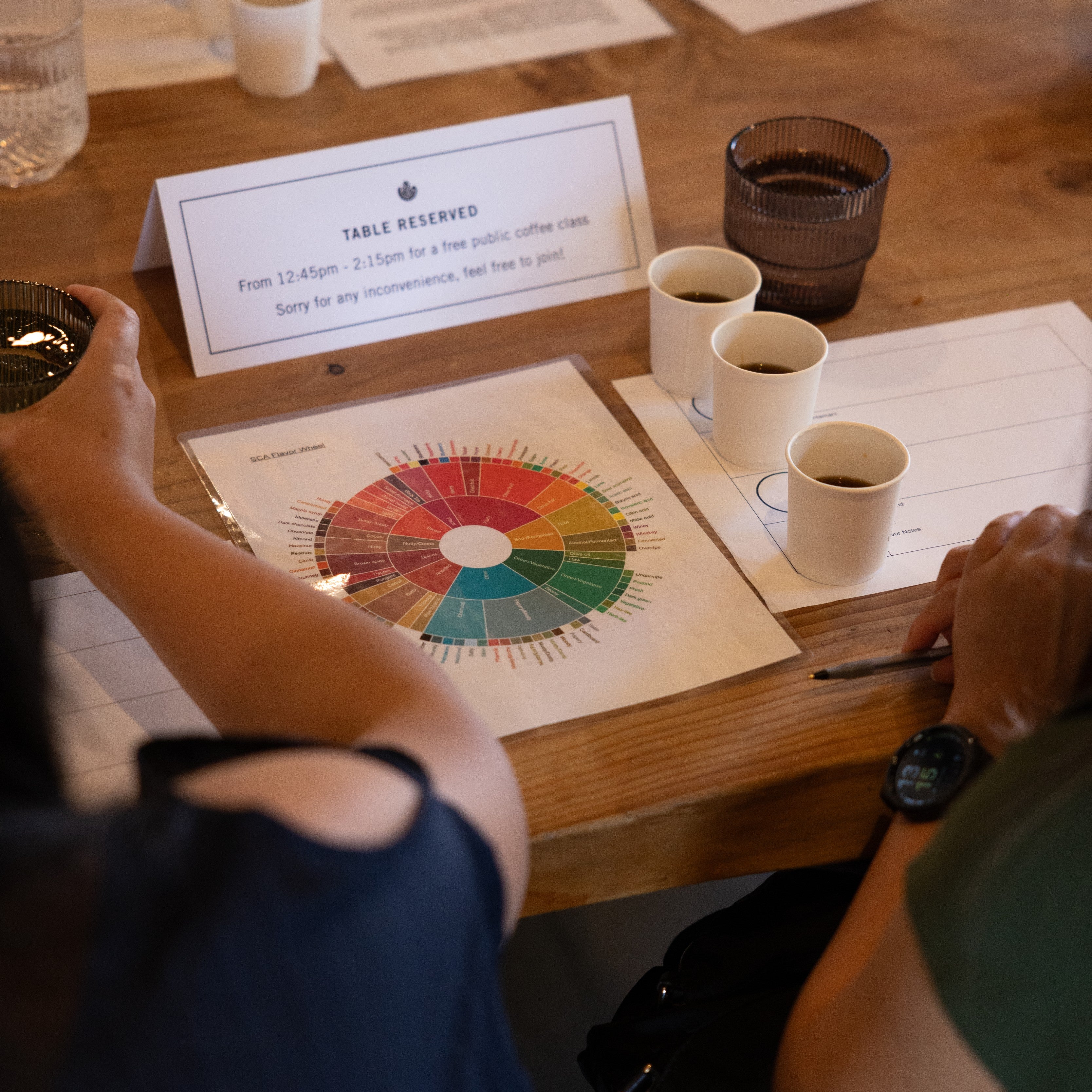 A wooden table at a coffee tasting event with several white espresso cups, printed color wheel cards, and a sign reading TABLE RESERVES. People are seated around the table in a real indoor setting, so this is Lifestyle rather than Product/Packaging.