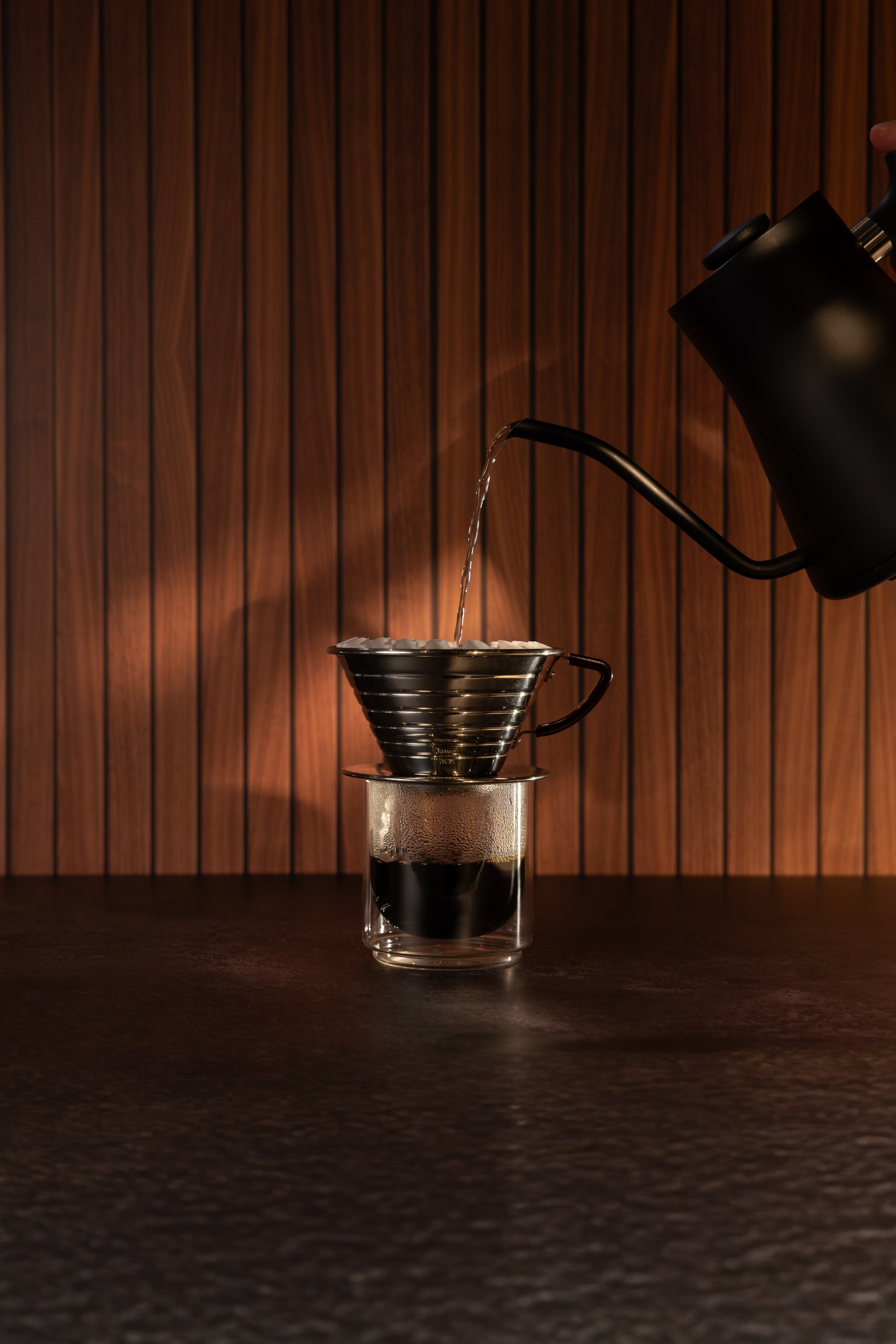 Pour-over coffee setup with a metal cone filter sitting on a glass pour-over cup, with hot water being poured from a matte black gooseneck kettle against a warm wood slat background.