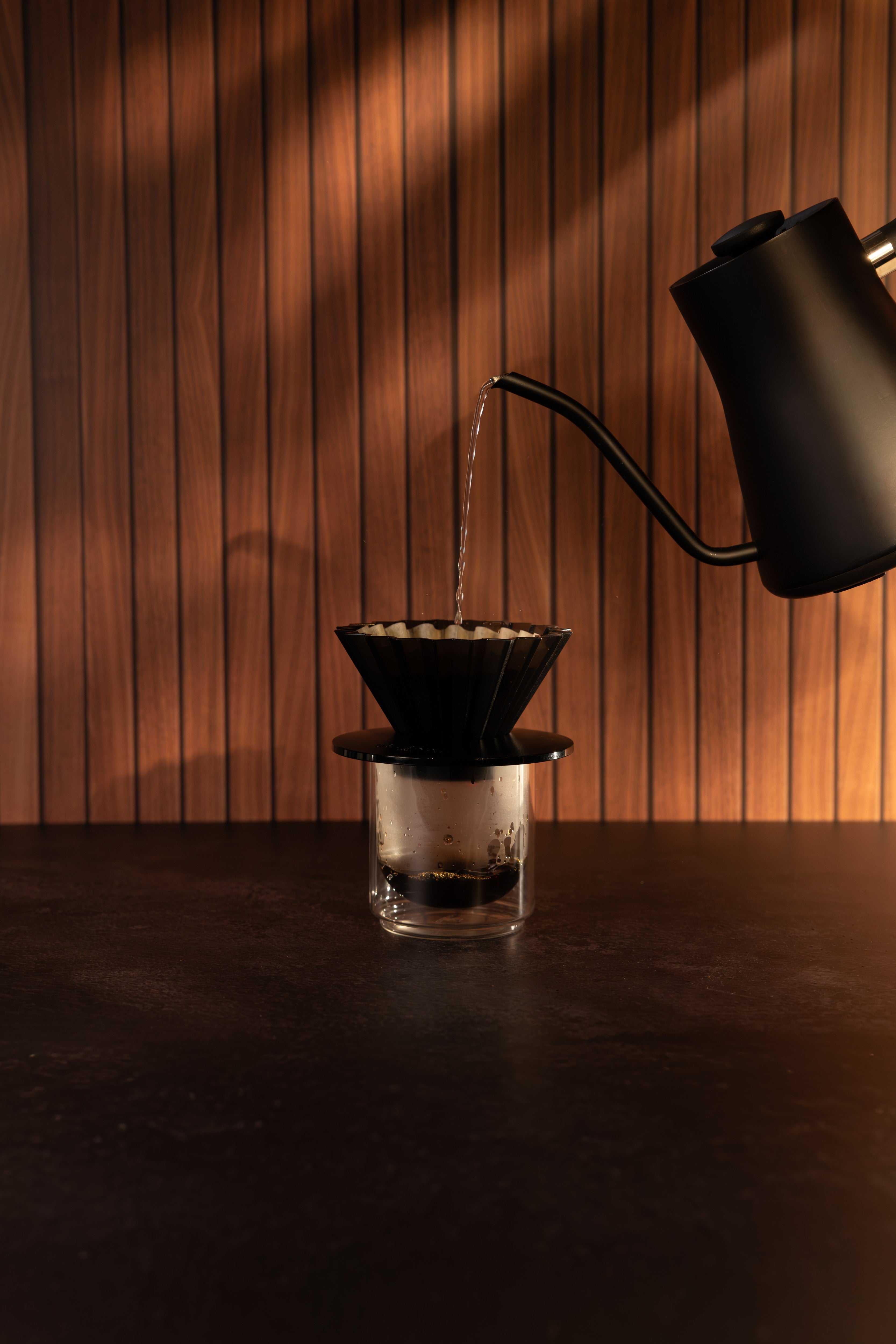 A pour-over coffee setup with a black cone filter and glass carafe on a dark table, with hot water being poured from a matte black gooseneck kettle against a warm wood-paneled background.