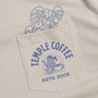 Close-up of a beige pocket on a shirt, featuring a blue 'Temple Coffee Estd. 2005' graphic and a blue monstera leaf print above it.