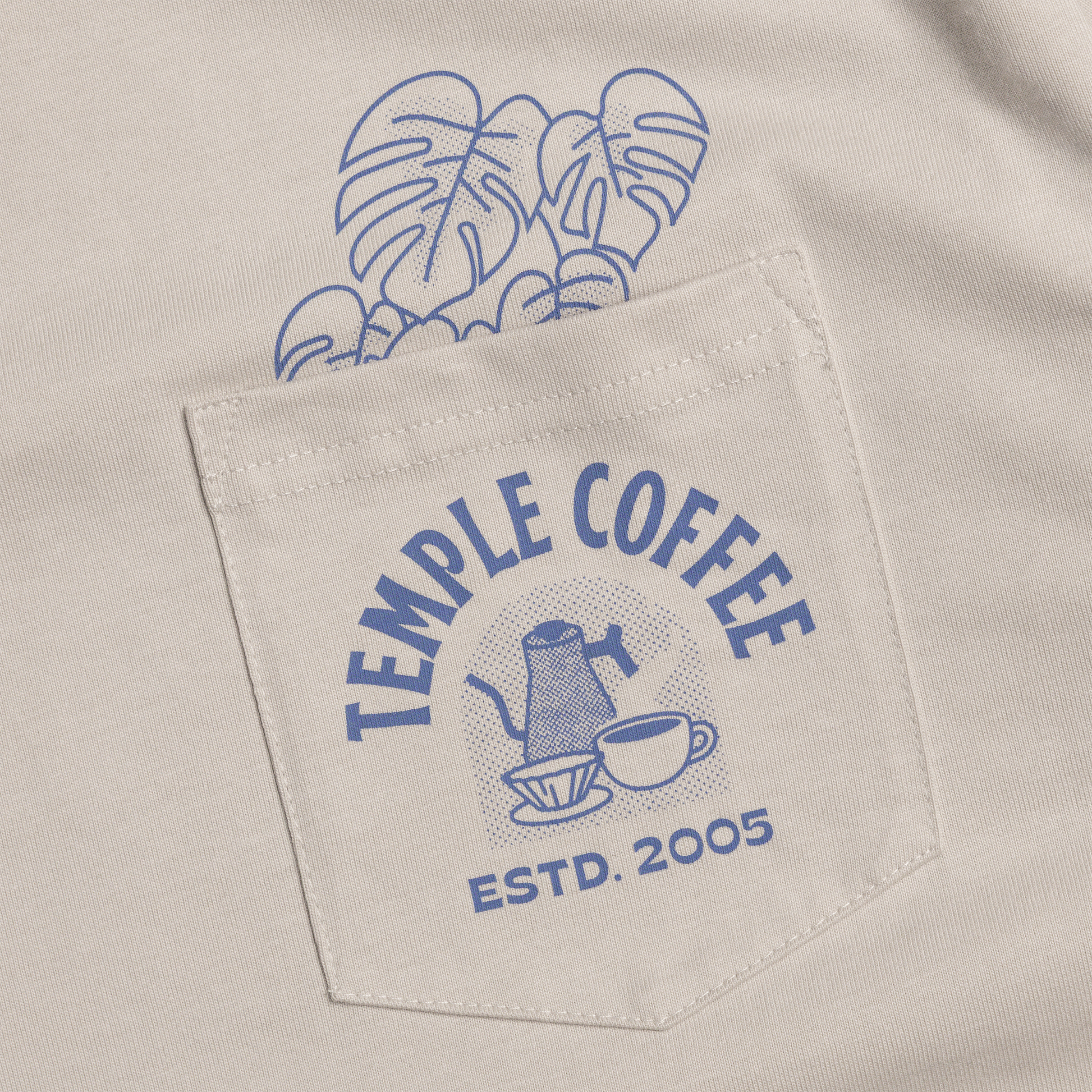 Close-up of a beige pocket on a shirt, featuring a blue 'Temple Coffee Estd. 2005' graphic and a blue monstera leaf print above it.
