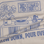 Close-up of a beige fabric product printed with a blue line-art coffee-themed illustration and text reading 'SLOW DOWN, POUR OVER' and 'BLOOMED'.