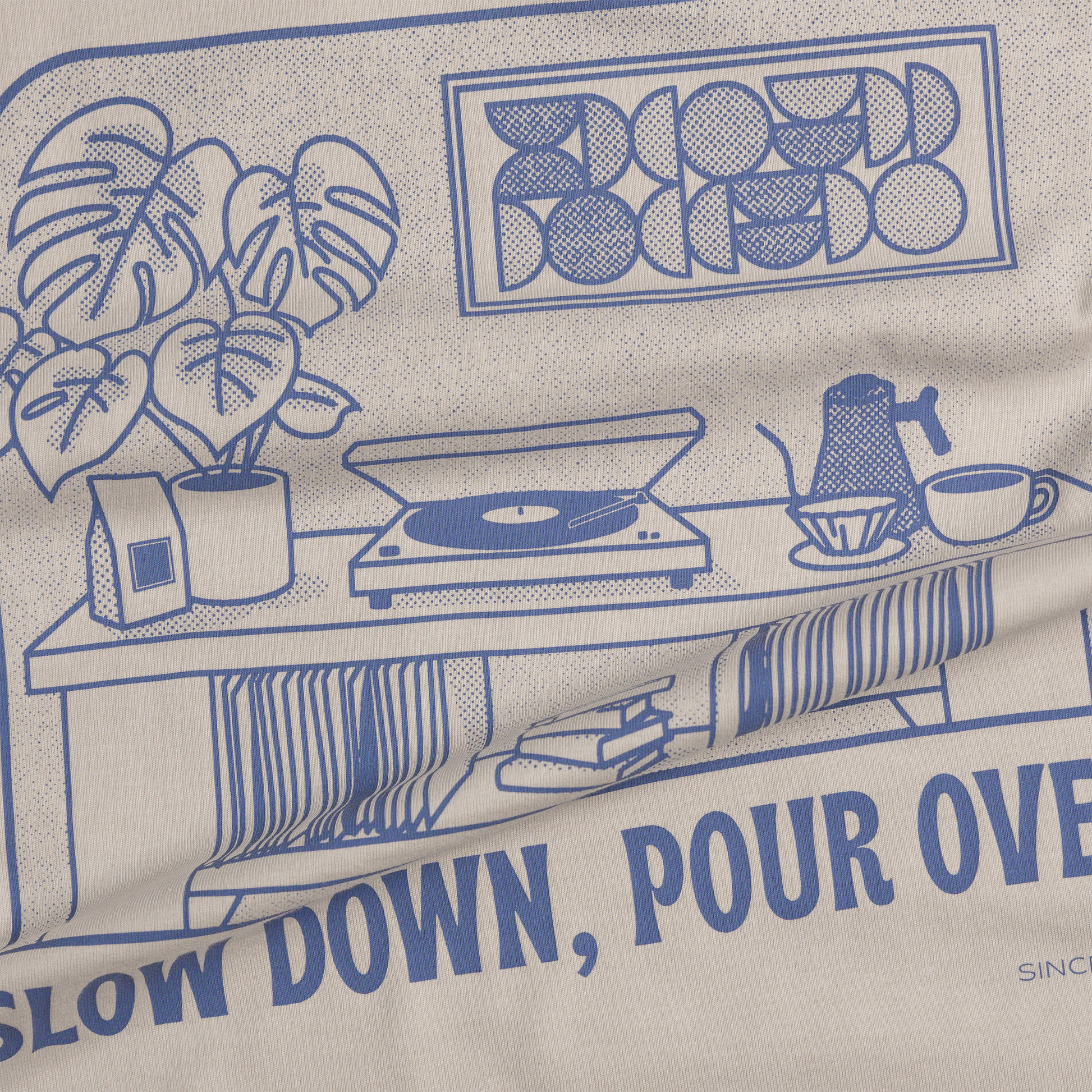 Close-up of a beige fabric product printed with a blue line-art coffee-themed illustration and text reading 'SLOW DOWN, POUR OVER' and 'BLOOMED'.