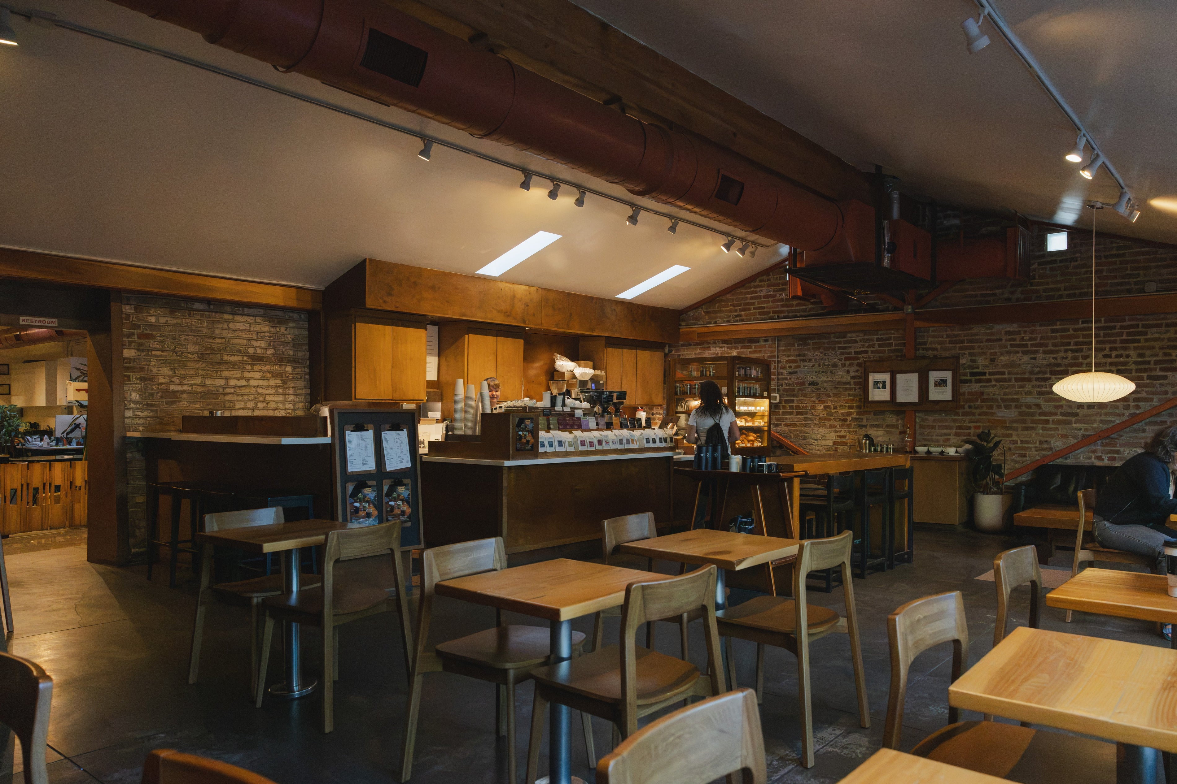 A cozy cafe interior with wooden tables and chairs, brick walls, exposed ceiling pipes, and a barista working behind the counter.